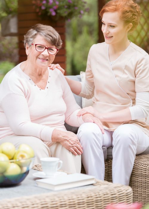 Tender professional caregiver in uniform putting her hand on a shoulder of an elderly woman during snack time on a patio of a senior home. Blurred background.